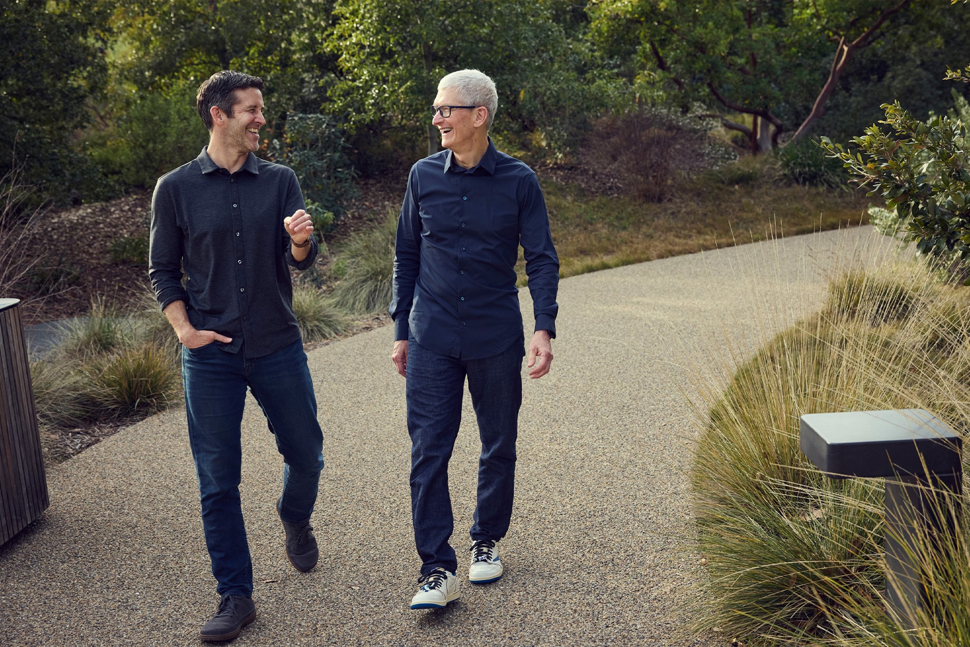 Two men walk side by side on a gravel path surrounded by greenery, smiling and engaged in conversation. Both are casually dressed in long-sleeve shirts, jeans, and sneakers.