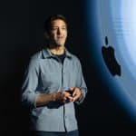 A man stands on stage speaking, holding a device, with a large Apple logo displayed on a screen behind him.