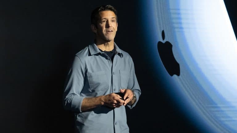 A man stands on stage speaking, holding a device, with a large Apple logo displayed on a screen behind him.