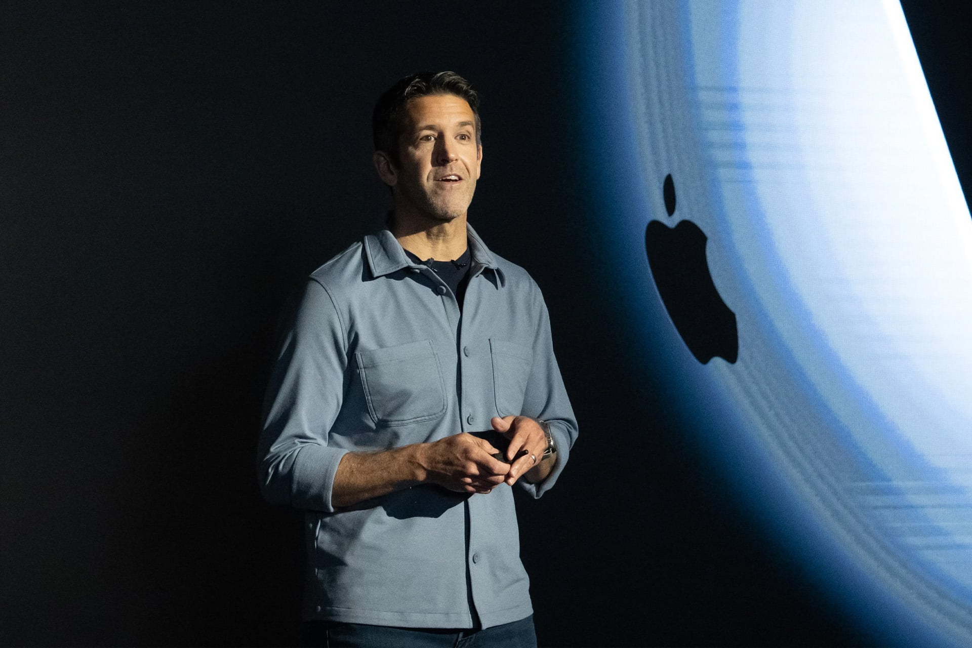 A man stands on stage speaking, holding a device, with a large Apple logo displayed on a screen behind him.