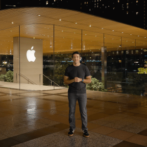 John Ternus stands smiling outside a modern glass Apple Store at night, with the illuminated Apple logo behind him and a tree on the right side of the image.