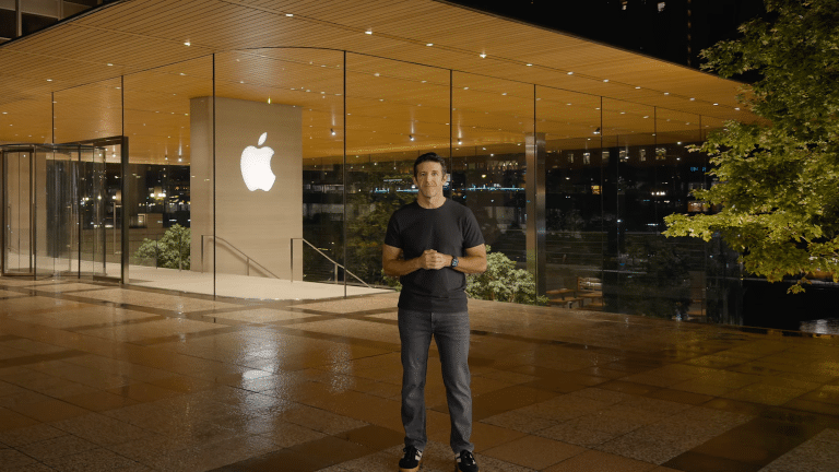 John Ternus stands smiling outside a modern glass Apple Store at night, with the illuminated Apple logo behind him and a tree on the right side of the image.