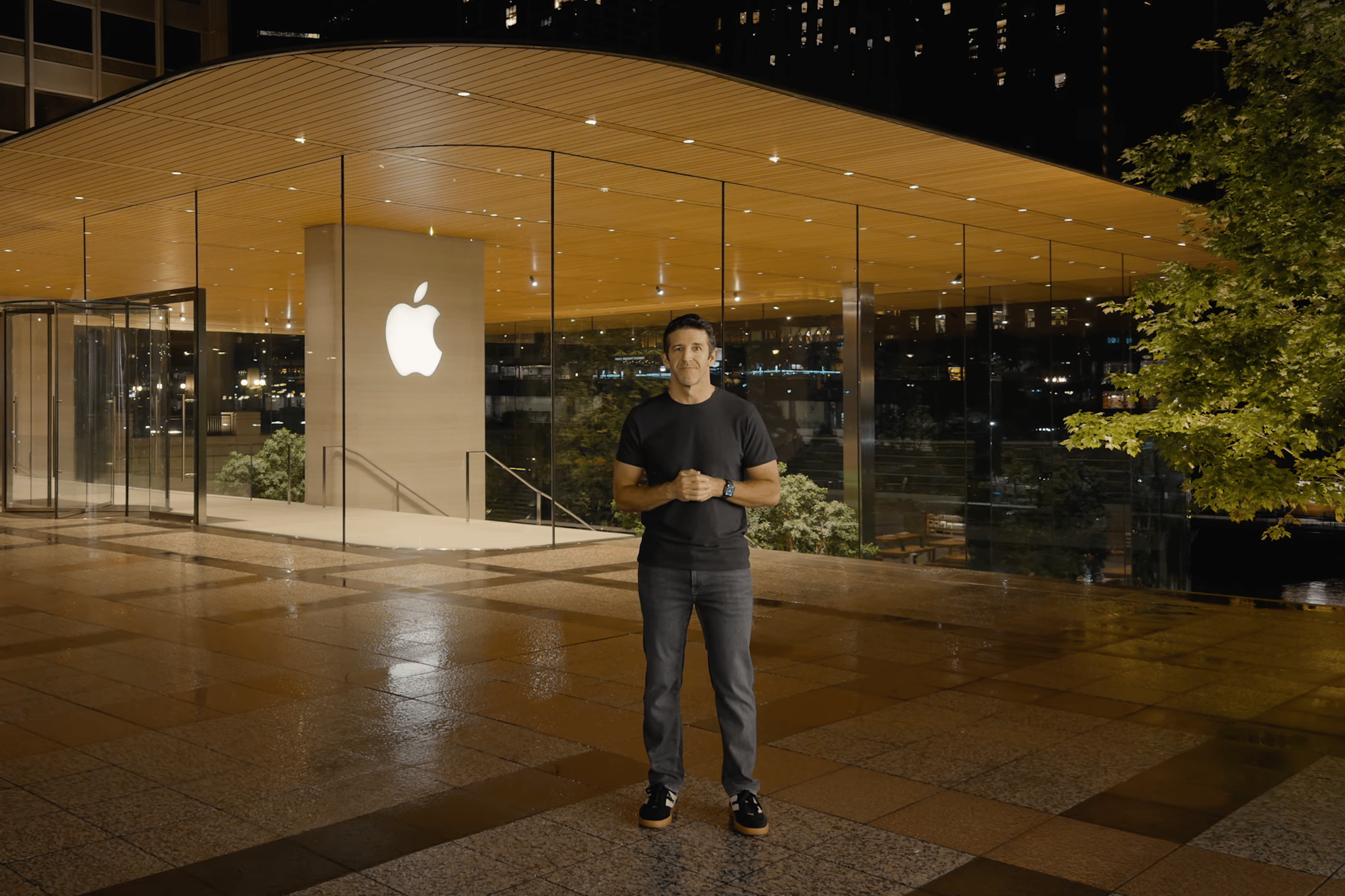 John Ternus stands smiling outside a modern glass Apple Store at night, with the illuminated Apple logo behind him and a tree on the right side of the image.