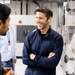 Three men stand in a modern industrial or laboratory setting. John Ternus, in a dark jacket, stands smiling with arms crossed, while another faces him, engaging in conversation. Large equipment is visible in the background.