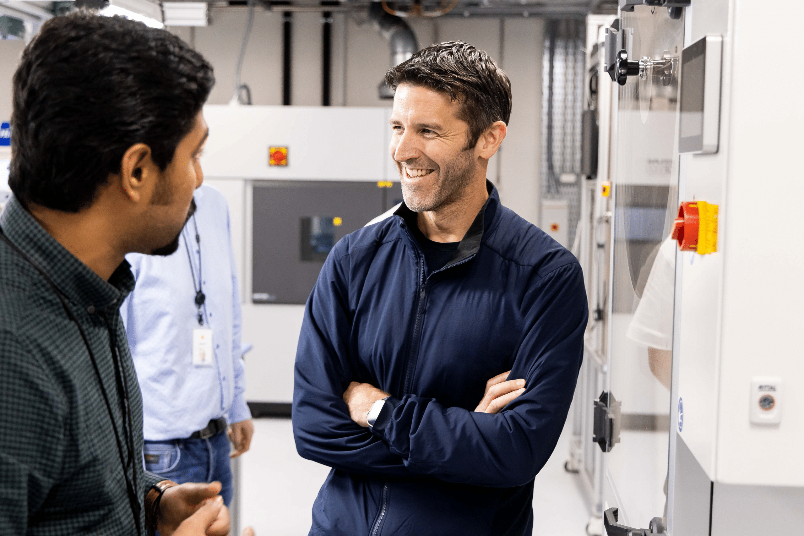 Three men stand in a modern industrial or laboratory setting. John Ternus, in a dark jacket, stands smiling with arms crossed, while another faces him, engaging in conversation. Large equipment is visible in the background.