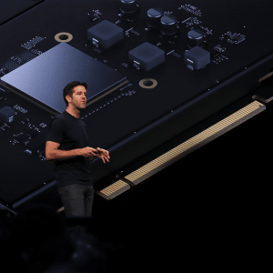 John Ternus, dressed in a black t-shirt and jeans, stands on stage delivering a presentation, with a large, detailed image of a computer chip and circuitry projected behind him.