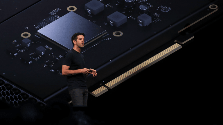 John Ternus, dressed in a black t-shirt and jeans, stands on stage delivering a presentation, with a large, detailed image of a computer chip and circuitry projected behind him.