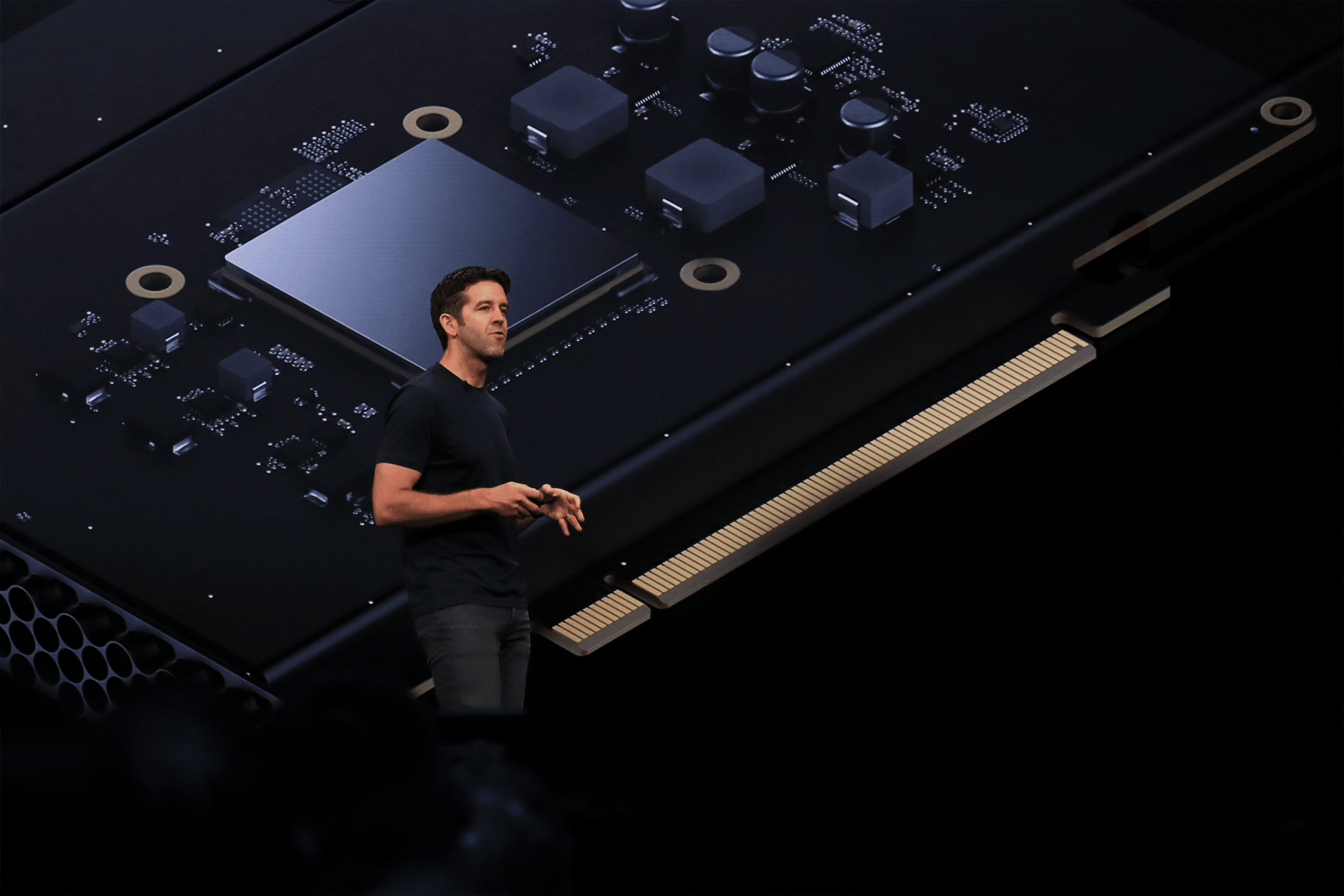John Ternus, dressed in a black t-shirt and jeans, stands on stage delivering a presentation, with a large, detailed image of a computer chip and circuitry projected behind him.