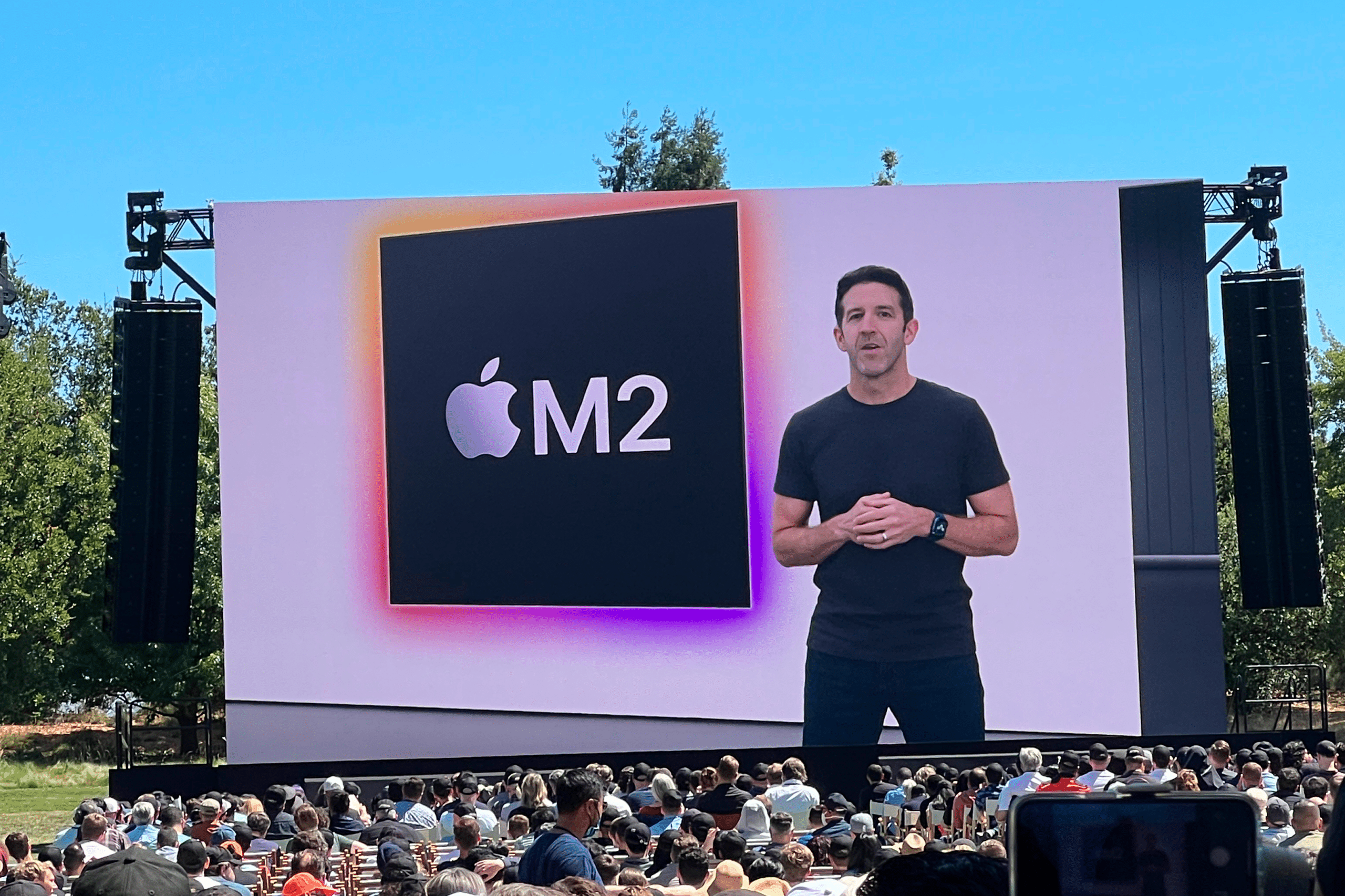 John Ternus presents on stage before a large outdoor audience, with a screen behind him displaying the Apple logo and “M2” in bold text. Trees and blue sky serve as the backdrop.