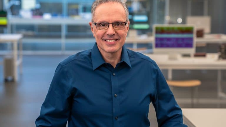 Johny Srouji, wearing glasses and a dark blue shirt, smiles while standing in a modern, brightly lit office or laboratory environment with desks and computer monitors in the background.