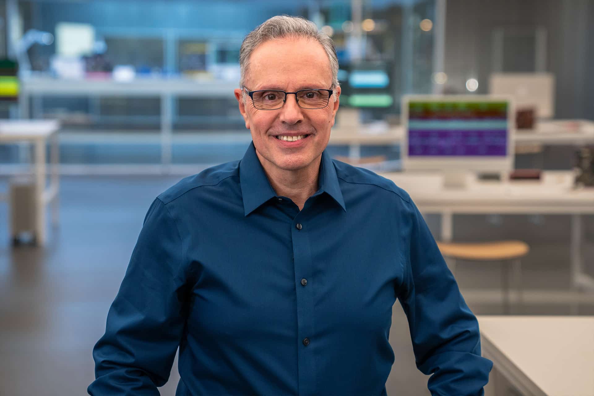 Johny Srouji, wearing glasses and a dark blue shirt, smiles while standing in a modern, brightly lit office or laboratory environment with desks and computer monitors in the background.