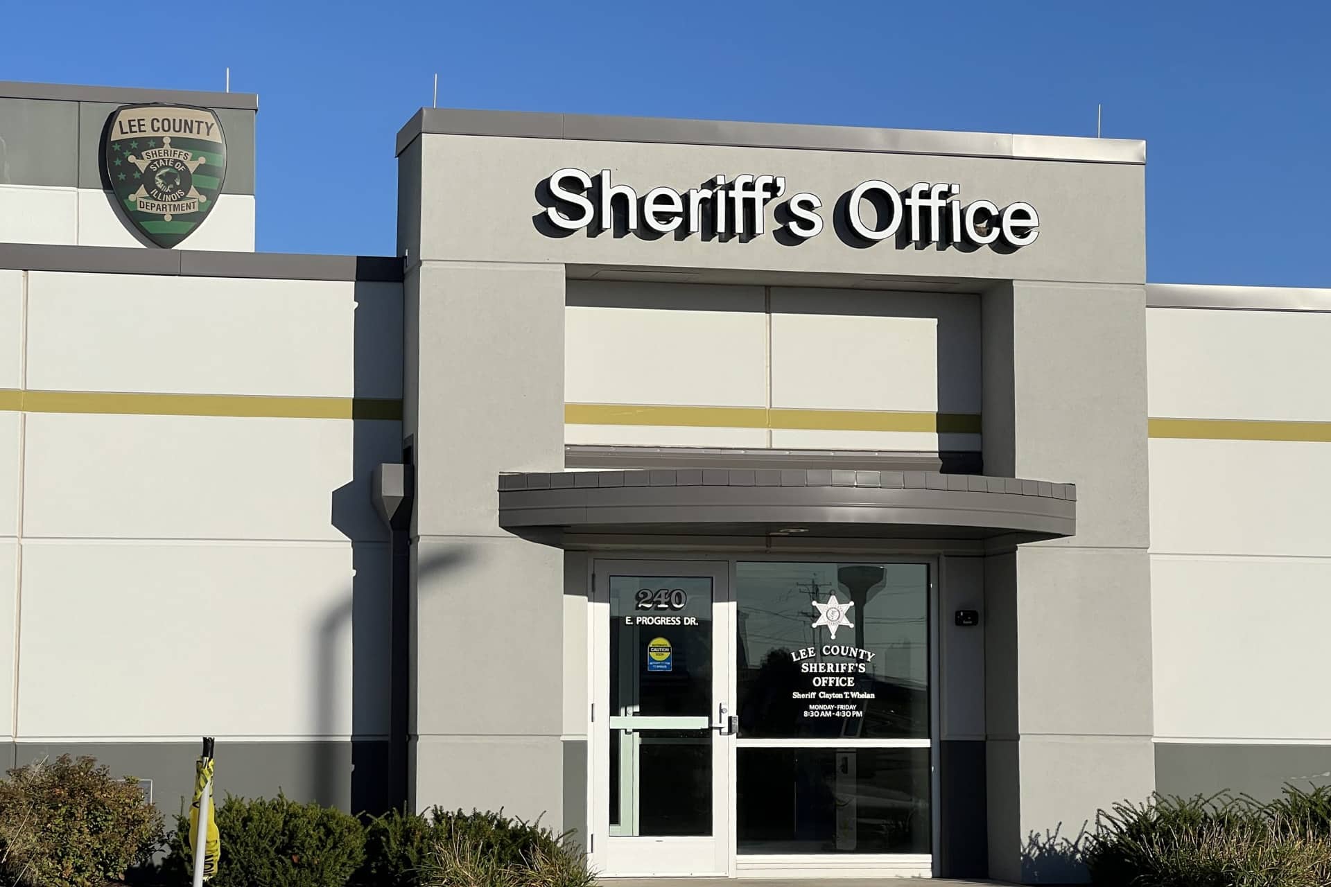 The front entrance of the Lee County Sheriff's Office building, with a large sign above the door and a badge logo on the window, stands ready to assist residents with concerns such as an Apple Account scam. The exterior is gray and white with trimmed bushes in front.