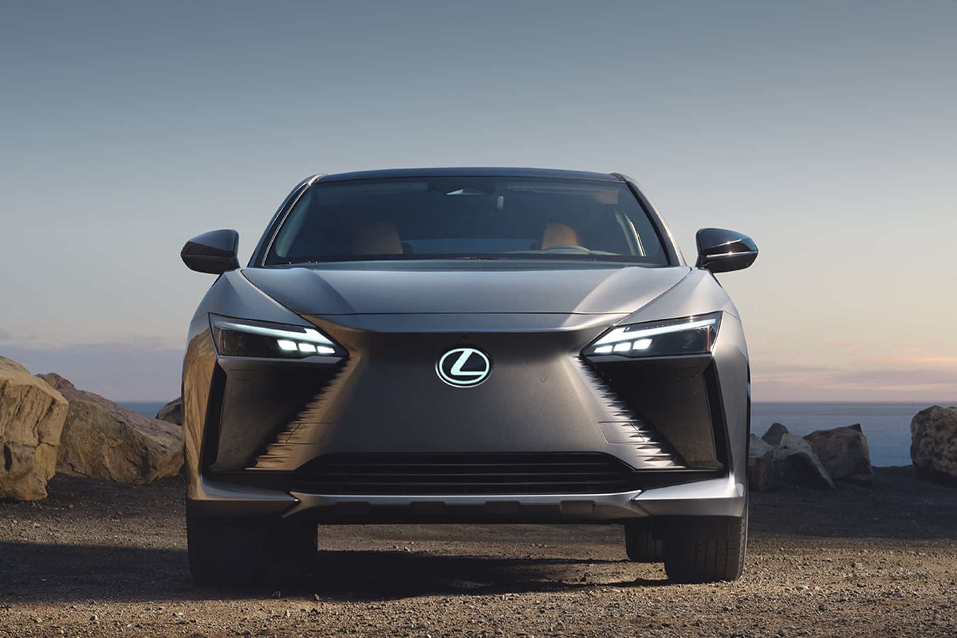 A front view of a silver Lexus electric vehicle parked outdoors on rocky terrain at sunset, its LED headlights shining and the Lexus logo on the grille—ready to be unlocked with your Lexus car key.