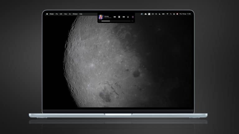 A MacBook displays a close-up photo of the moon's surface as its wallpaper, with a small music player widget at the top center. The minimalist dark gray background adds to the Magic Island feel of this celestial workspace.