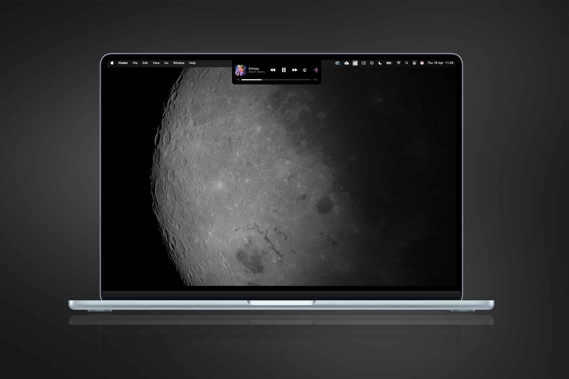 A MacBook displays a close-up photo of the moon's surface as its wallpaper, with a small music player widget at the top center. The minimalist dark gray background adds to the Magic Island feel of this celestial workspace.