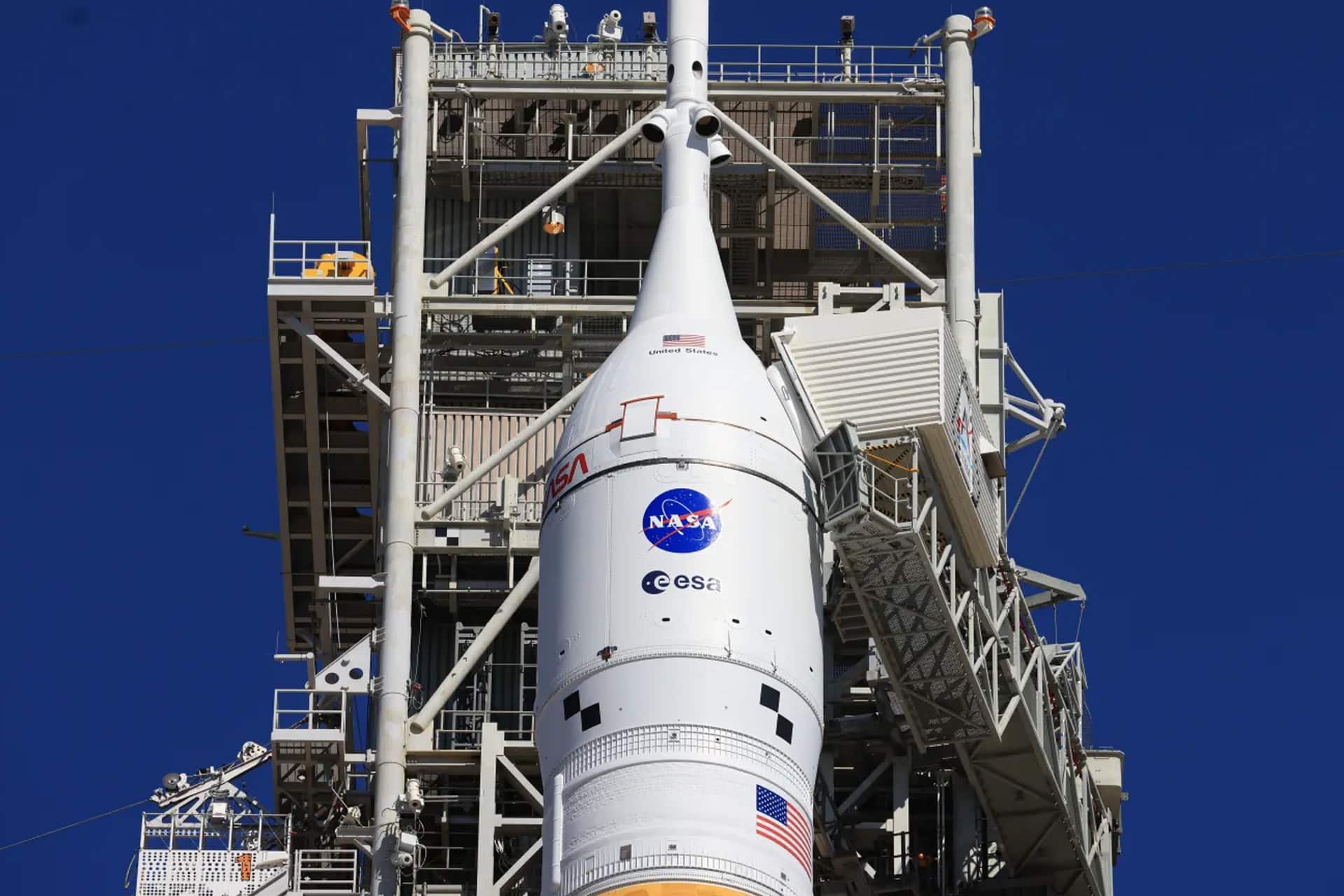 A close-up view of a NASA rocket, featuring Artemis II and ESA logos on its exterior, stands vertically at the launch pad under a clear blue sky—captured sharply as if seen through the latest iPhones.