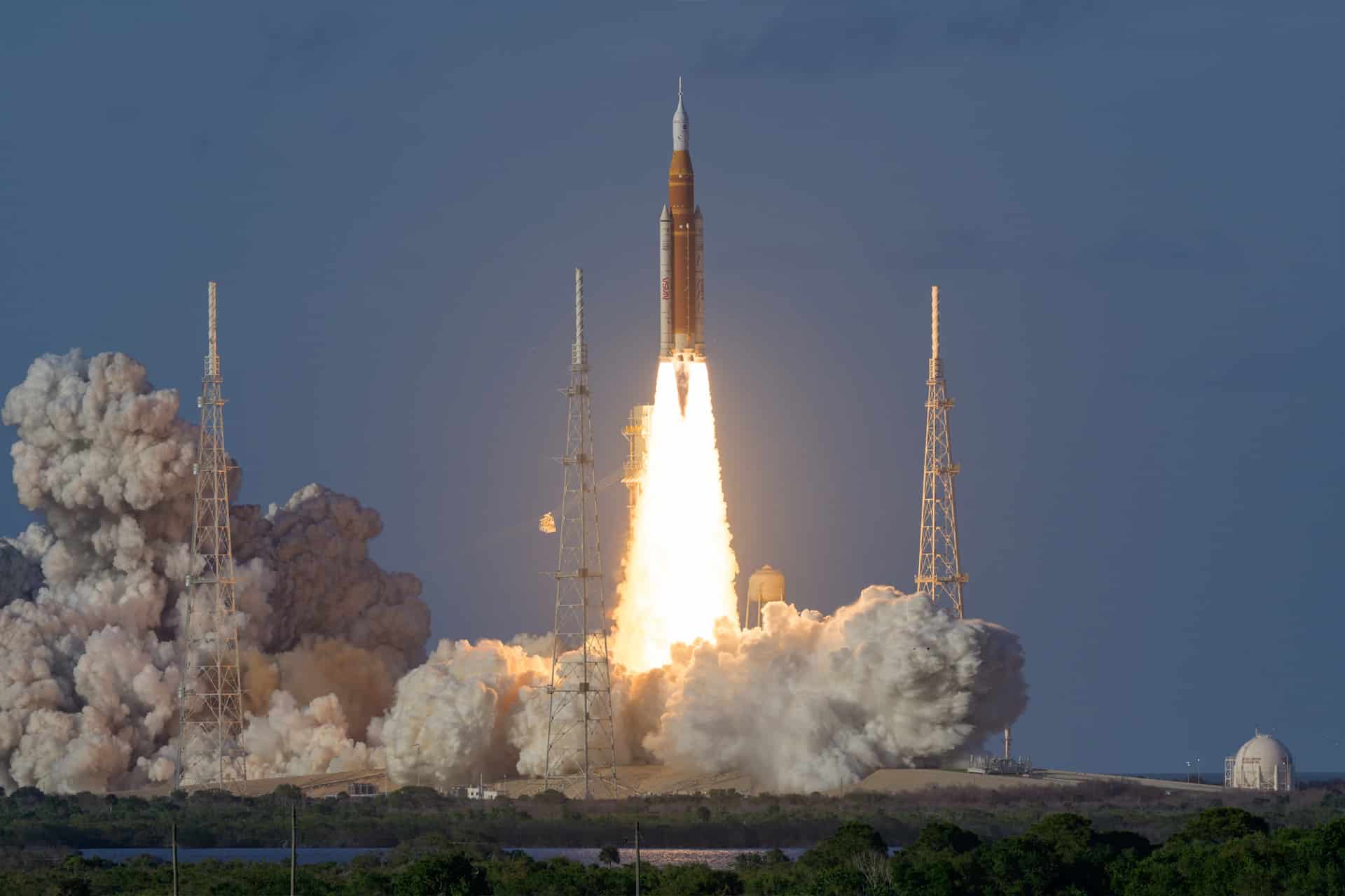 A rocket, reminiscent of the Artemis II mission, lifts off from its launch pad, producing bright flames and thick clouds of smoke, with a clear sky and launch towers visible in the background.