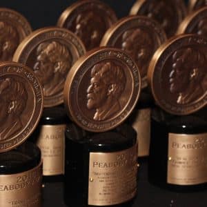 Rows of Peabody Awards from the 86th Annual Peabody Awards, featuring bronze medallions with a profile portrait, are displayed on black bases with gold nameplates, arranged closely together against a dark background.