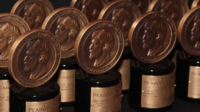 Rows of Peabody Awards from the 86th Annual Peabody Awards, featuring bronze medallions with a profile portrait, are displayed on black bases with gold nameplates, arranged closely together against a dark background.