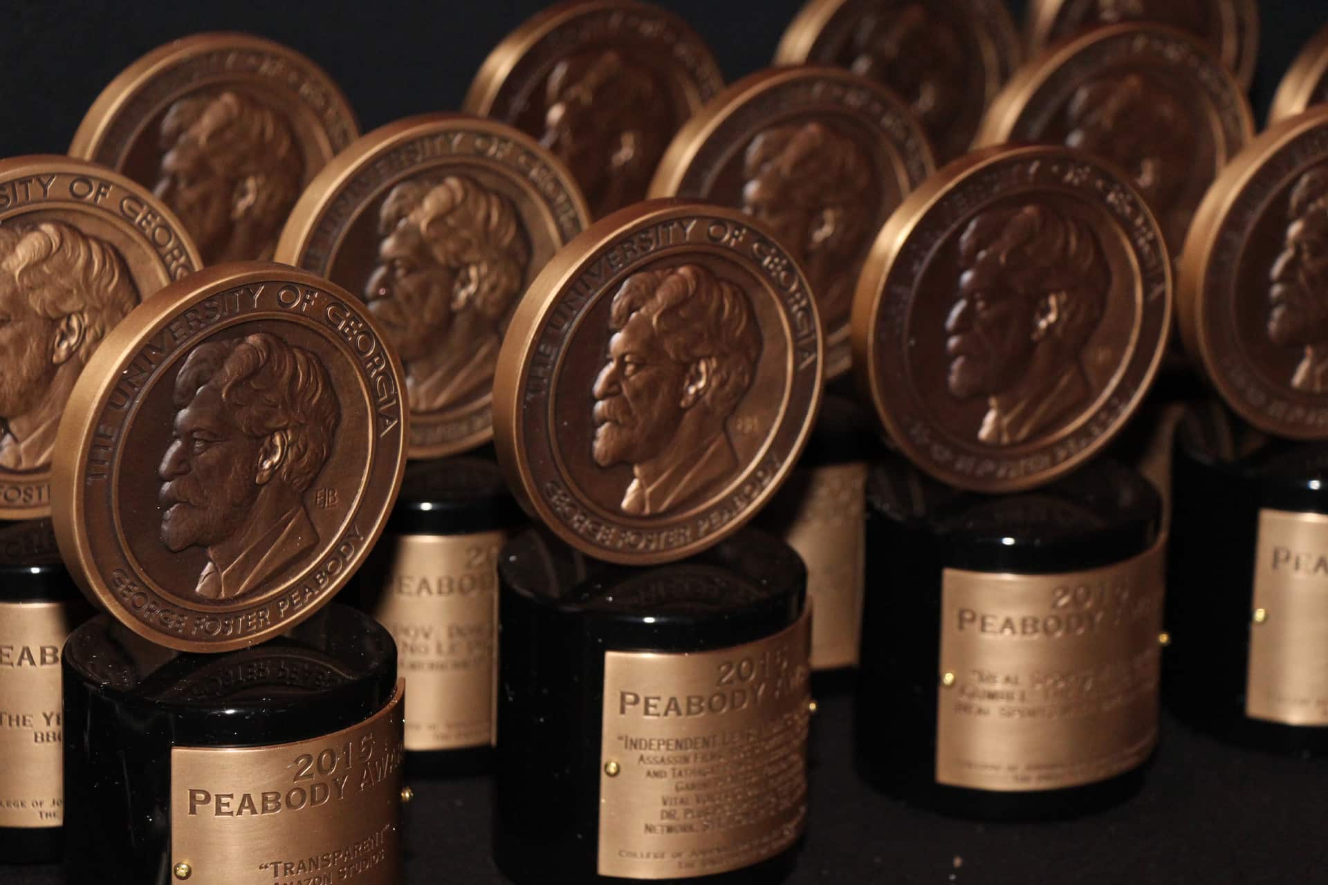 Rows of Peabody Awards from the 86th Annual Peabody Awards, featuring bronze medallions with a profile portrait, are displayed on black bases with gold nameplates, arranged closely together against a dark background.