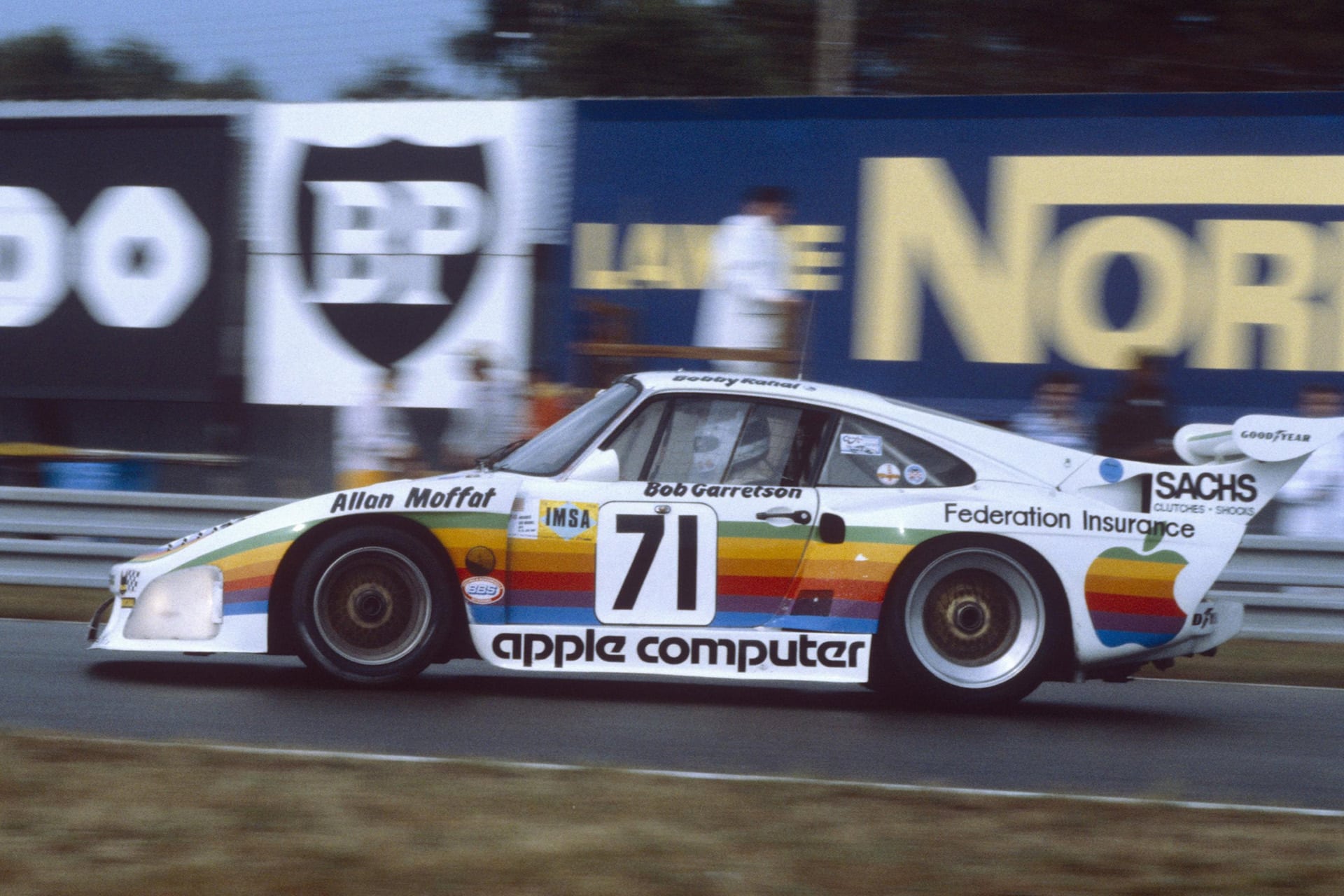 A vintage Porsche Apple race car with rainbow Apple Computer branding and the number 71 speeds down the racetrack, passing large advertisement boards. The car features sponsor logos and a distinctive rear wing.