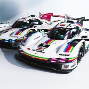 Two white Porsche Apple race cars with rainbow stripes and retro “Apple Computer” logos are parked side by side in a studio setting, proudly displaying racing numbers and various sponsor logos on their sleek bodies.