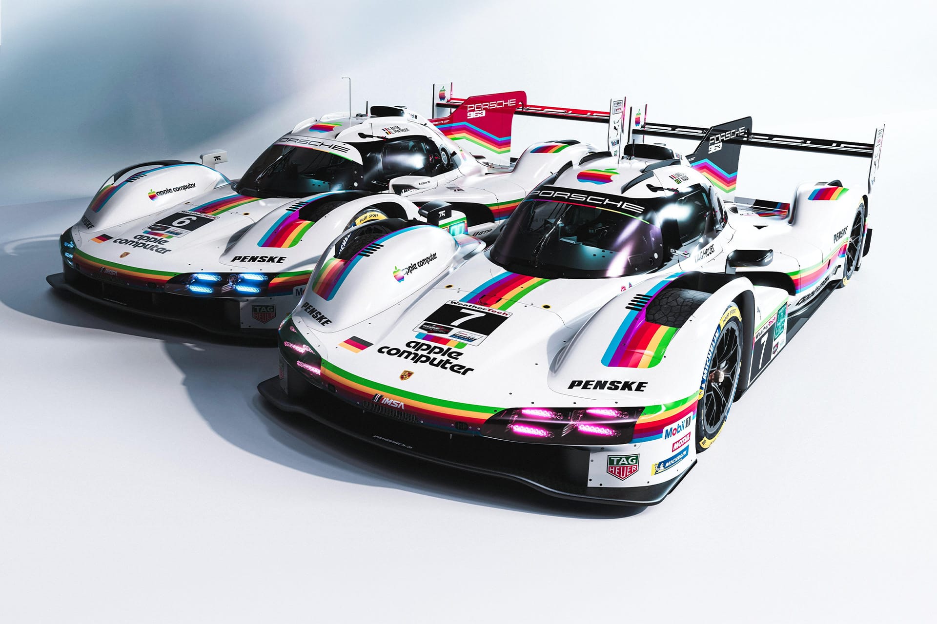 Two white Porsche Apple race cars with rainbow stripes and retro “Apple Computer” logos are parked side by side in a studio setting, proudly displaying racing numbers and various sponsor logos on their sleek bodies.