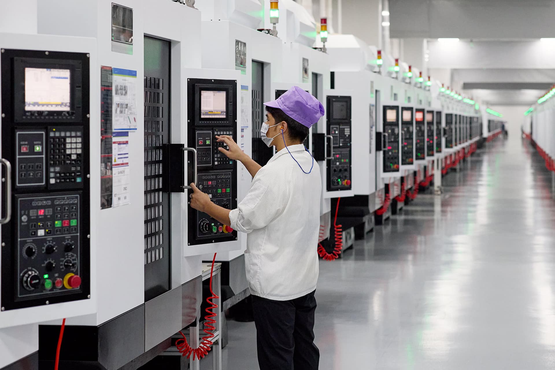 A worker in a white uniform and purple cap operates a control panel on a CNC machine, possibly shaping Apple rare earth components, in a brightly lit, clean factory with rows of similar machines in the background.