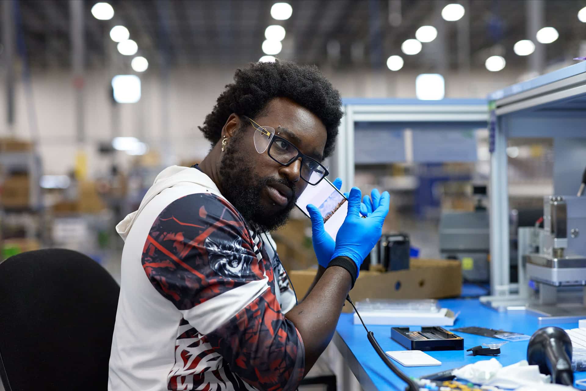 A man wearing glasses and blue gloves sits at a workstation in a factory, holding up a small Apple rare earth electronic device and looking at the camera. He is surrounded by tools and equipment on the desk.