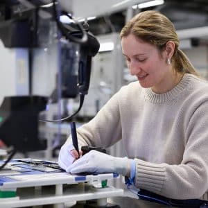 A woman wearing gloves and a light sweater uses a tool to work on Apple rare earth electronic components at a workstation in a modern lab or manufacturing facility.