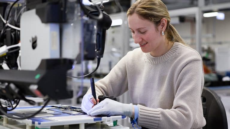 A woman wearing gloves and a light sweater uses a tool to work on Apple rare earth electronic components at a workstation in a modern lab or manufacturing facility.