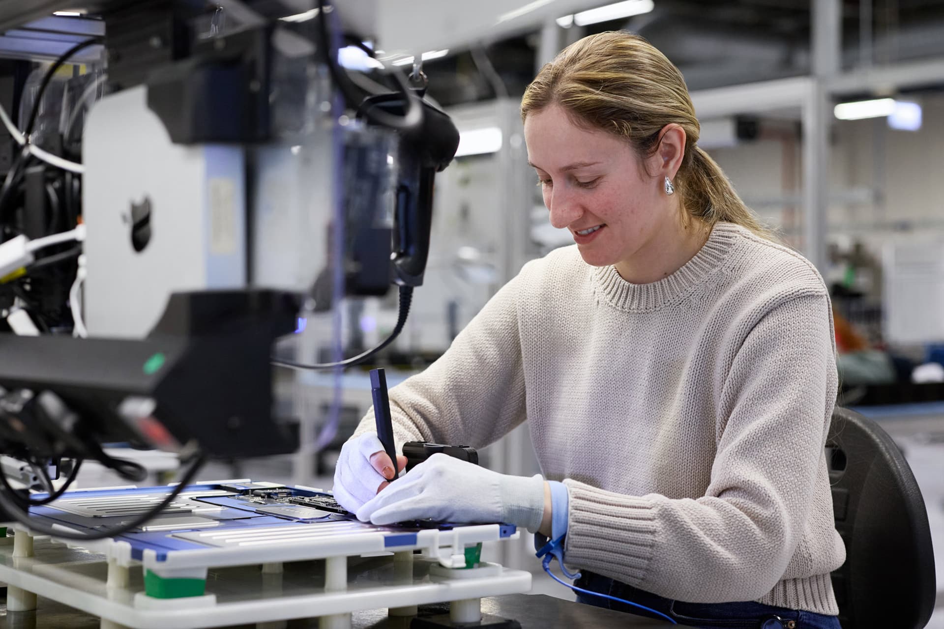 A woman wearing gloves and a light sweater uses a tool to work on Apple rare earth electronic components at a workstation in a modern lab or manufacturing facility.