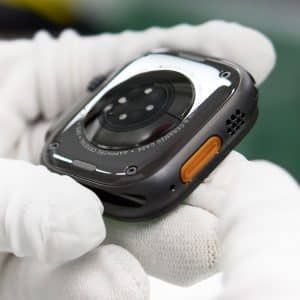 A gloved hand holds the back of a smartwatch, showing its sensors, an orange side button, part of its metal casing, and hints of Apple rare earth materials used in its construction.