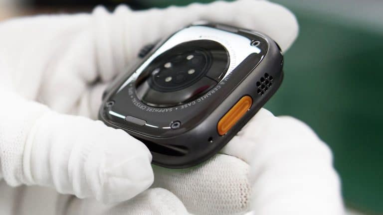 A gloved hand holds the back of a smartwatch, showing its sensors, an orange side button, part of its metal casing, and hints of Apple rare earth materials used in its construction.
