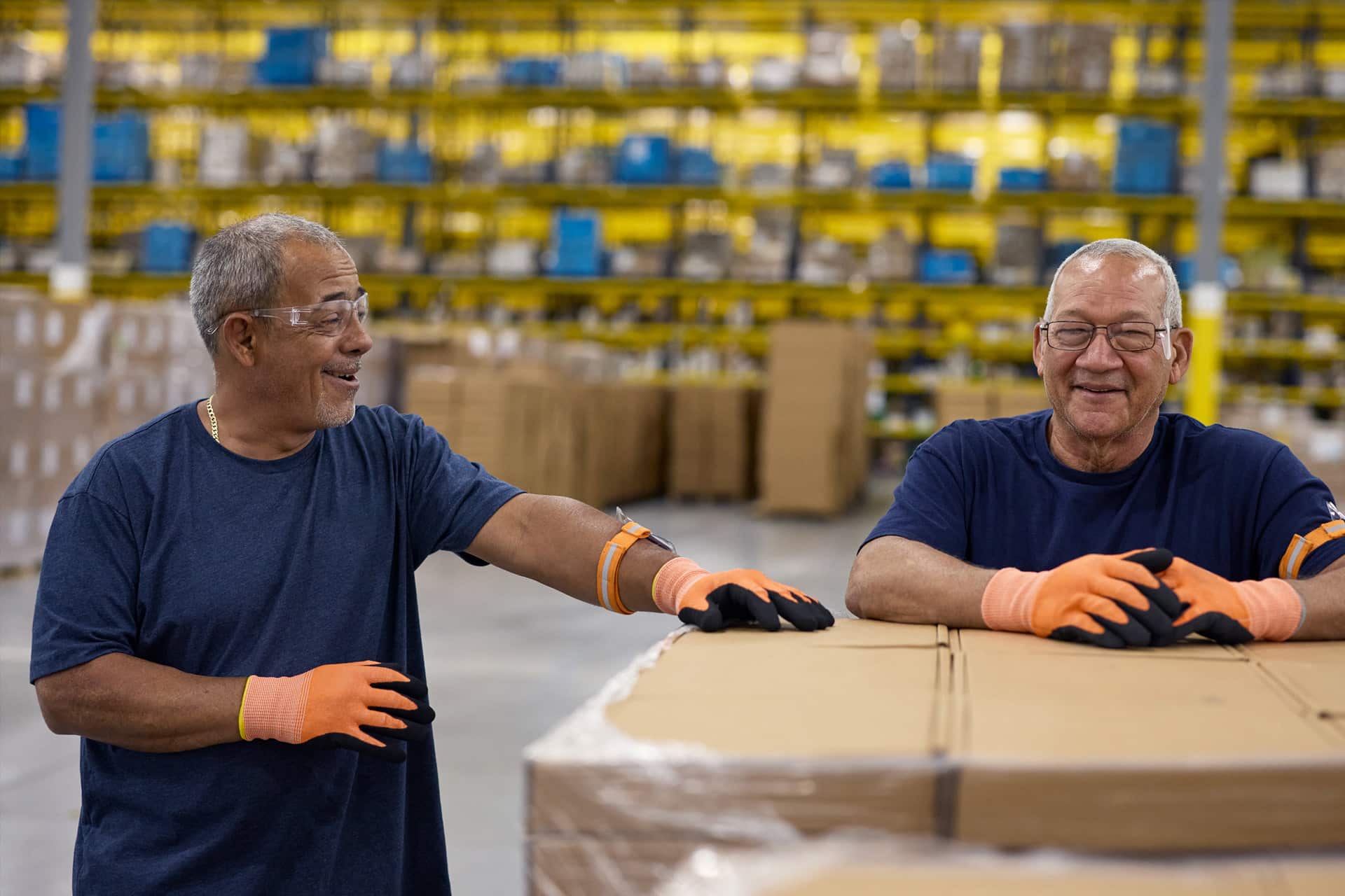 Two older men wearing navy shirts, safety glasses, and orange gloves smile and talk while leaning on a large pallet of Apple rare earth boxes in a warehouse with shelves full of products in the background.