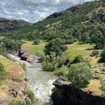 A river flows through a rocky canyon surrounded by green hills and trees under a partly cloudy sky. Sunlight highlights the vibrant landscape, reminiscent of nature’s beauty inspiring Apple recycled materials in sustainable innovation.
