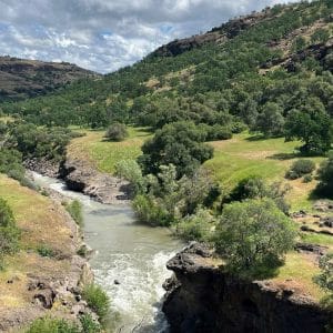 A river flows through a rocky canyon surrounded by green hills and trees under a partly cloudy sky. Sunlight highlights the vibrant landscape, reminiscent of nature’s beauty inspiring Apple recycled materials in sustainable innovation.