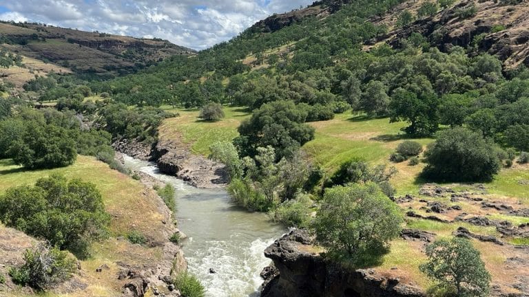A river flows through a rocky canyon surrounded by green hills and trees under a partly cloudy sky. Sunlight highlights the vibrant landscape, reminiscent of nature’s beauty inspiring Apple recycled materials in sustainable innovation.