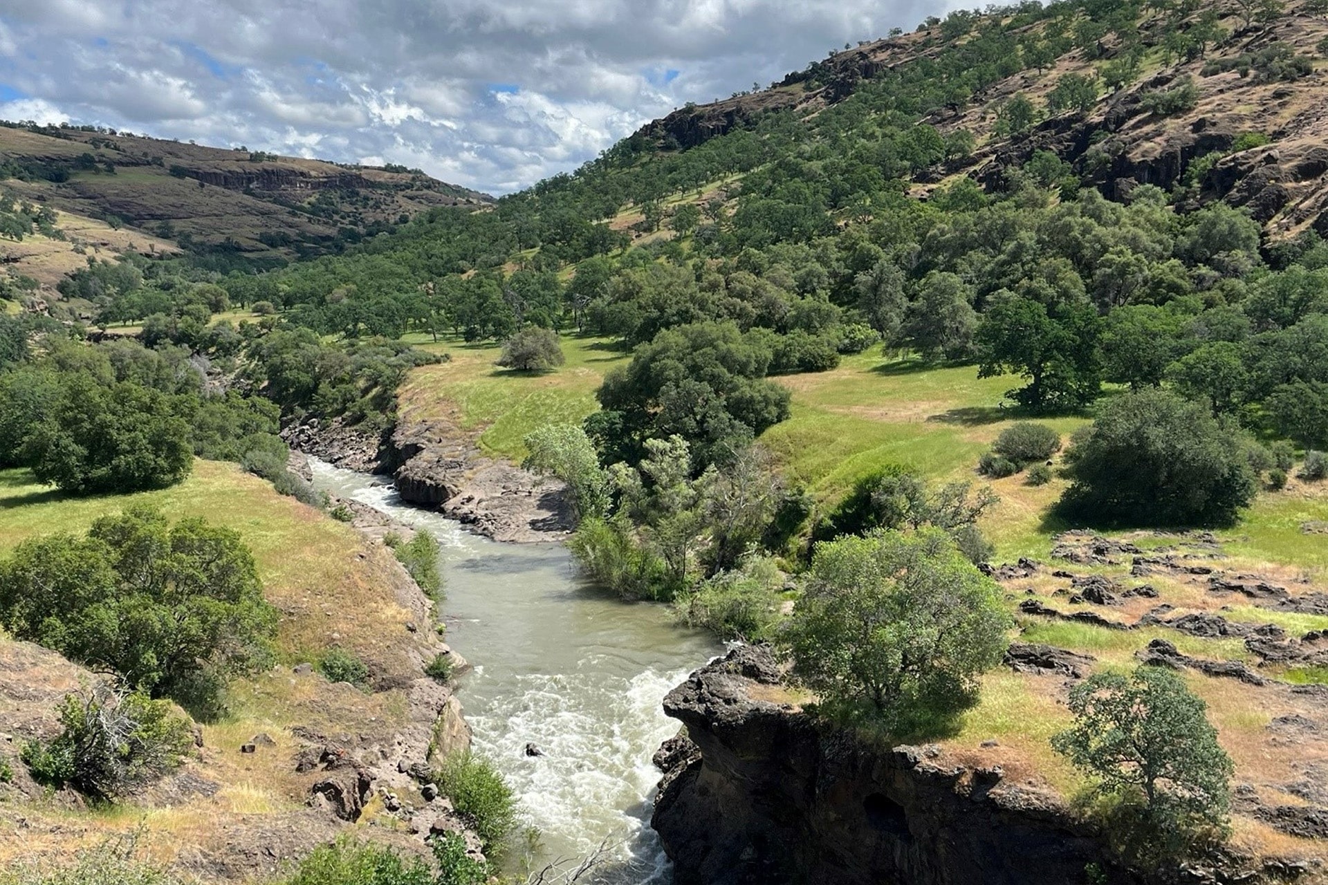 A river flows through a rocky canyon surrounded by green hills and trees under a partly cloudy sky. Sunlight highlights the vibrant landscape, reminiscent of nature’s beauty inspiring Apple recycled materials in sustainable innovation.
