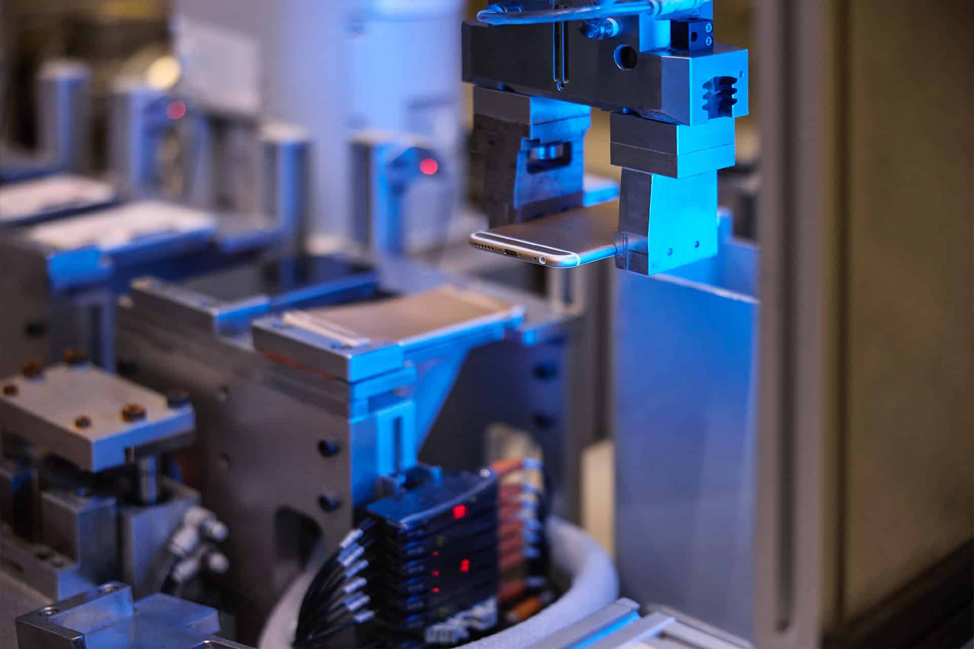 A robotic arm with a metal gripper is handling a slim, rectangular electronic device made from Apple recycled materials in an automated manufacturing environment with various machinery and components visible.