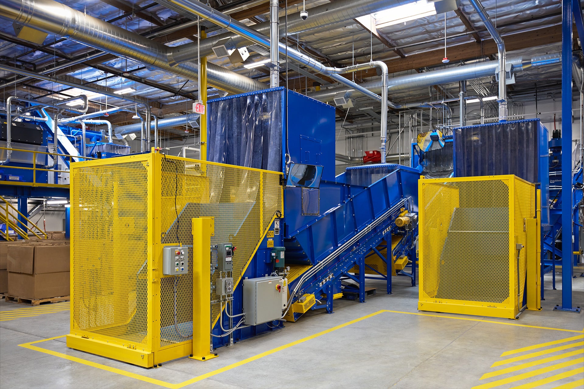 A modern recycling facility interior with conveyor belts, blue machinery, and yellow safety barriers. Overhead pipes and lights are visible, along with control panels and a few cardboard boxes made from Apple recycled materials on the floor.