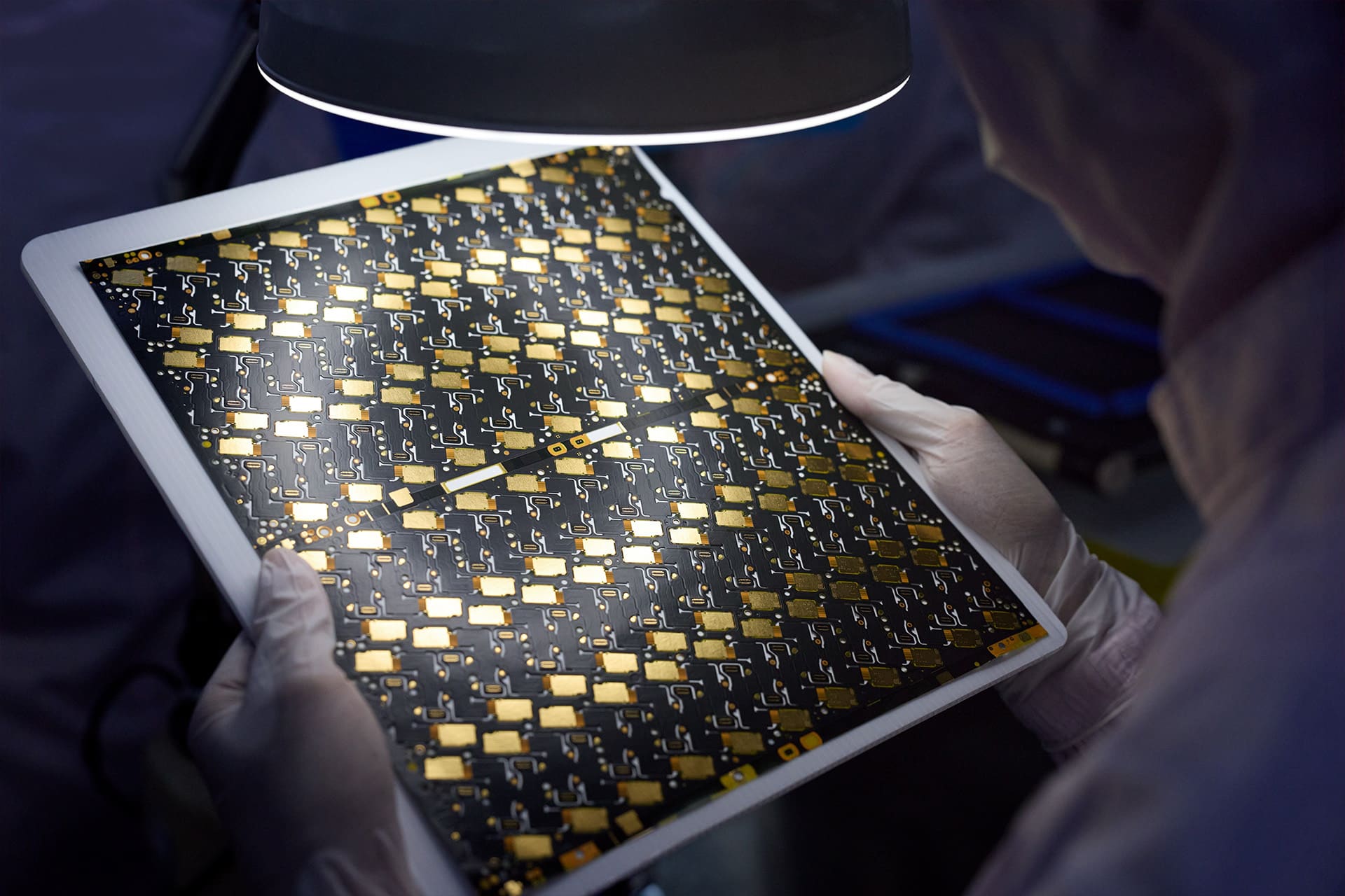 A person wearing gloves and protective clothing inspects a large semiconductor wafer—crafted with Apple recycled materials and featuring intricate gold and black circuit patterns—under a bright lamp in a cleanroom environment.