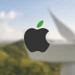 The black Apple logo with a green leaf is centered over a blurred background of a wind turbine and green hills, symbolizing Apple sustainability partners' commitment to renewable energy and environmental sustainability.