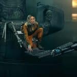 A person in an orange jumpsuit crouches on a damaged metal bridge suspended over a dark, industrial setting, evoking the tense atmosphere of Silo Season 3 with crumbling structures and dim lighting in the background.