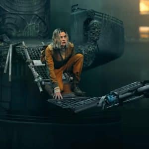 A person in an orange jumpsuit crouches on a damaged metal bridge suspended over a dark, industrial setting, evoking the tense atmosphere of Silo Season 3 with crumbling structures and dim lighting in the background.