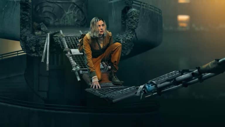 A person in an orange jumpsuit crouches on a damaged metal bridge suspended over a dark, industrial setting, evoking the tense atmosphere of Silo Season 3 with crumbling structures and dim lighting in the background.