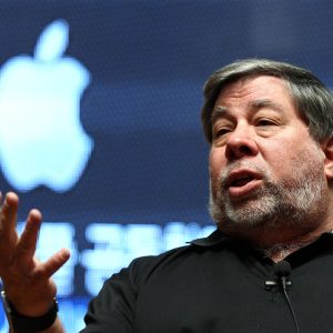 A man with gray hair and a beard speaks animatedly while gesturing with his hand. He wears a black shirt and sits in front of a large, illuminated Apple logo, resembling Steve Wozniak during an engaging talk.