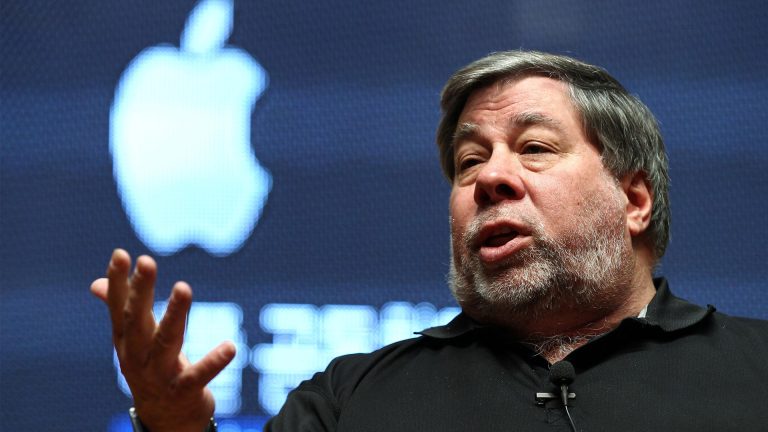 A man with gray hair and a beard speaks animatedly while gesturing with his hand. He wears a black shirt and sits in front of a large, illuminated Apple logo, resembling Steve Wozniak during an engaging talk.