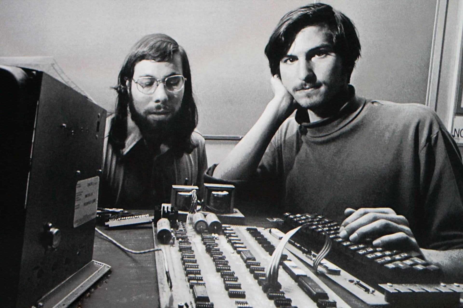 Two young men sit at a table with an early computer circuit board and electronic components in a black-and-white photo. One, Steve Wozniak, has glasses and a beard; the other is clean-shaven, resting his head on his hand. Both look toward the camera.