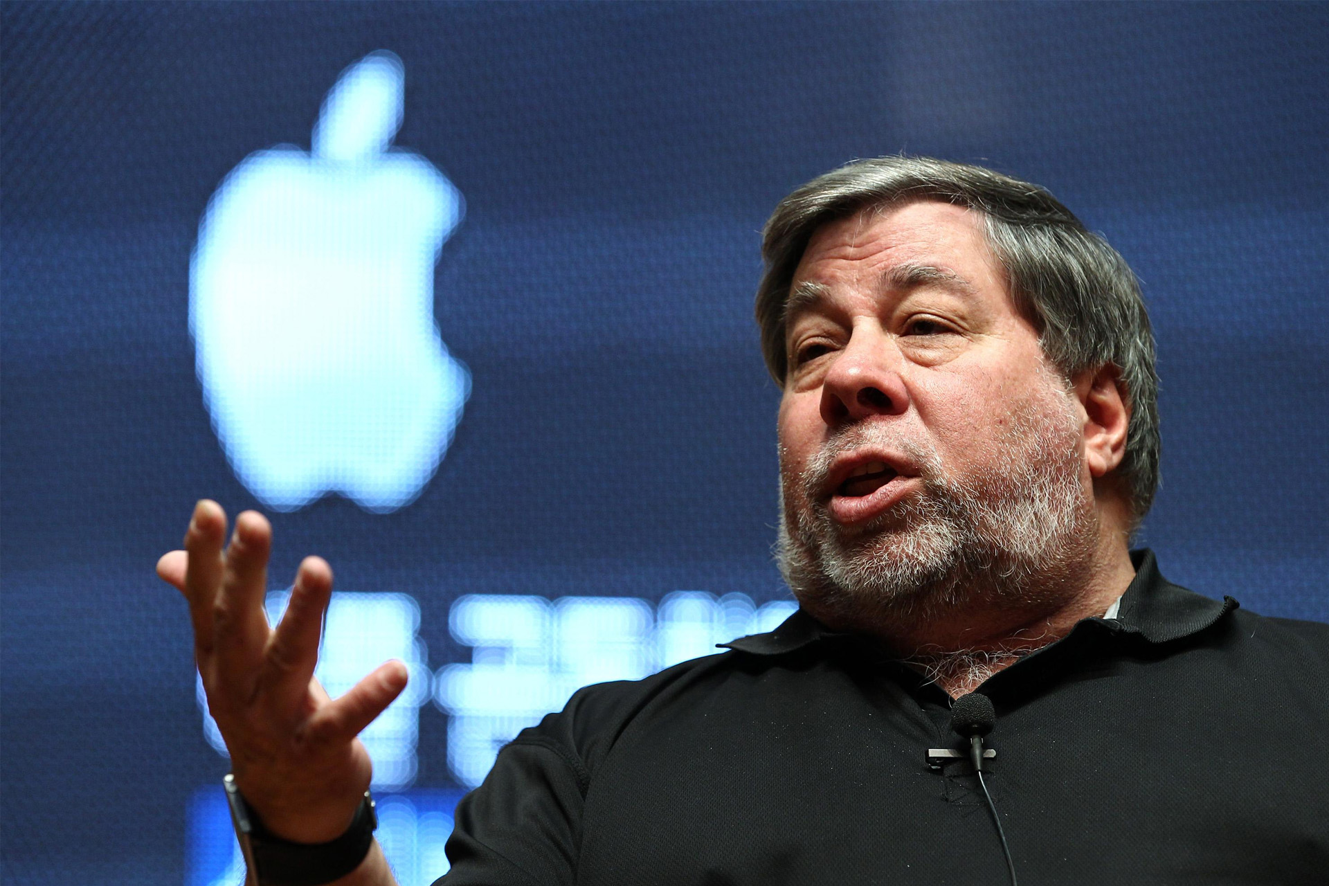 A man with gray hair and a beard speaks animatedly while gesturing with his hand. He wears a black shirt and sits in front of a large, illuminated Apple logo, resembling Steve Wozniak during an engaging talk.